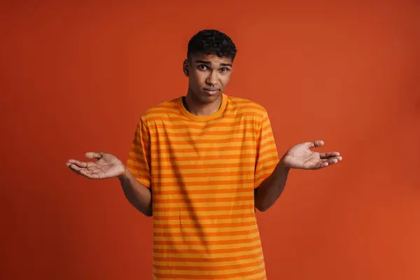 Young handsome confused african man spreading hands to the sides and looking ar camera, while standing over isolated orange background