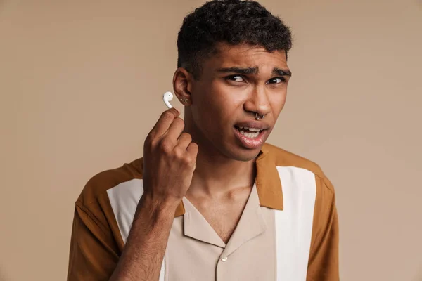 年轻英俊 困惑的非洲男子穿着无线耳机的画像 从耳朵里掏出来 看着相机 站在孤立的米黄色背景上 — 图库照片