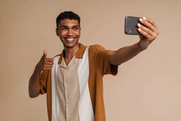 年轻的非洲帅哥面带微笑 拿着手机自拍 摆出大拇指的样子 站在孤零零的米色背景上 — 图库照片