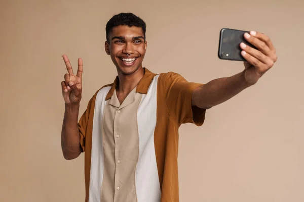 年轻的非洲帅哥面带微笑 一边拿着手机自拍 摆出胜利的架势 一边站在孤立的米色背景上 — 图库照片