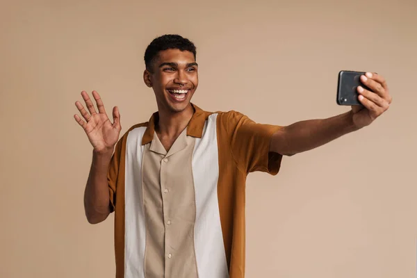 年轻的非洲帅哥面带微笑 一边拿着手机和挥手自拍 一边站在孤立的米黄色背景上 — 图库照片