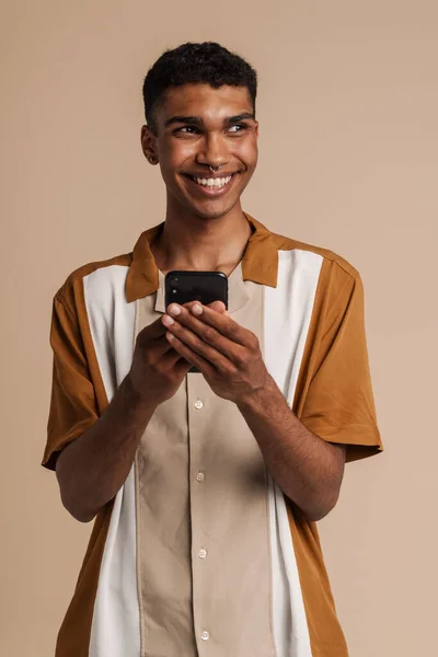 年轻的非洲帅哥面带微笑 一边拿着电话 一边看一边站在孤零零的米色背景上 — 图库照片