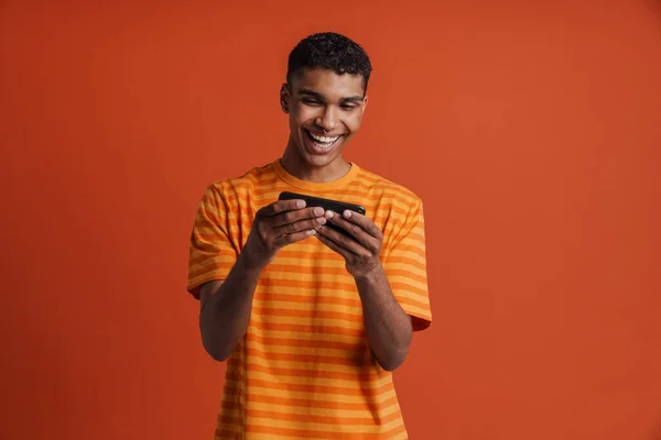 年轻英俊的笑脸快乐的非洲男人一边在手机上玩游戏 一边站在孤立的橙色背景下 — 图库照片