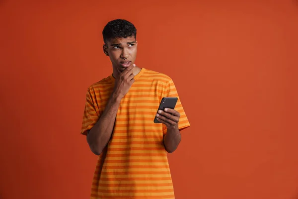 年轻英俊 体贴的非洲男人 一边拿着电话 一边看一边摸着下巴 一边站在孤立的橙色背景上 — 图库照片