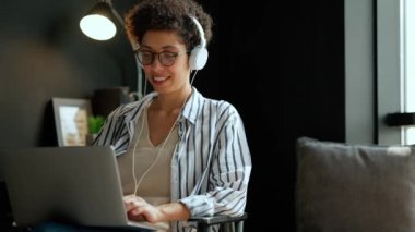 Gülümseyen kıvırcık saçlı kadın evde kulaklıkla dizüstü bilgisayarda çalışıyor.