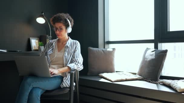 可爱的西班牙卷发女人 戴着耳机在笔记本电脑上工作 看着家里的窗户 — 图库视频影像