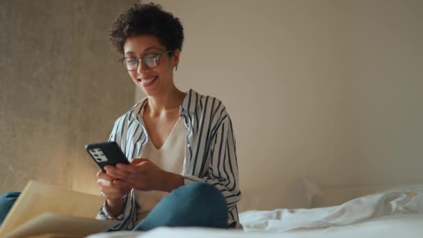 快乐的西班牙卷发女人在手机上发短信 床上放着一本书 — 图库视频影像