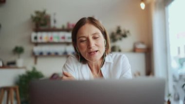 Beyaz tişörtlü mutlu yaşlı kadın kafedeki laptopta kulaklıkla video konuşmalarıyla konuşuyor.