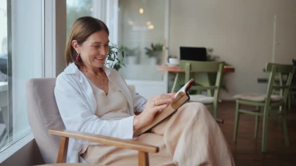 カフェに座っている間に白いシャツの読書本で高齢者の女性を楽しむ — ストック動画