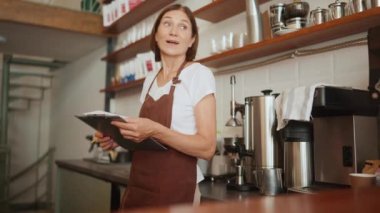 Kâğıt döküman yazan ve kafede tezgahın arkasına bakan mutlu, olgun bir bayan barista.