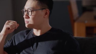 Gözlüklü düşünceli Asyalı adam evde otururken yanına bakıyor.