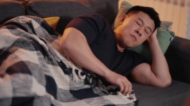 Yorgun Asyalı adam evde kanepede uzanırken televizyon seyrediyor ve esniyor.