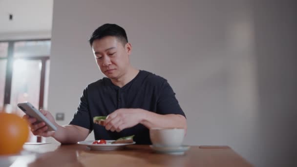 Düşünceli Asyalı Adam Sandviç Yiyor Evde Kahvaltıda Telefonla Mesaj Atıyor — Stok video