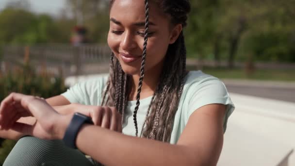 ワークアウト後に屋外で腕時計に刺繍のピグテールテキストを持つリラックスしたアフリカの女性 スポーツ活動 アクティブなライフスタイル — ストック動画