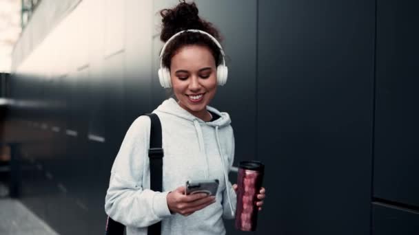ワークアウトの後に屋外に歩いている間 携帯電話を入力してヘッドフォンでスポーティーな女性を笑顔 スポーツ活動 アクティブなライフスタイル — ストック動画