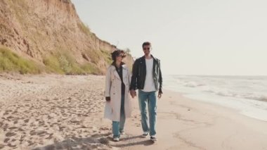 Mutlu çift deniz kıyısında yürürken konuşuyor ve el ele tutuşuyor.