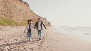Mutlu çift el ele tutuşup deniz kıyısında koşuyor.