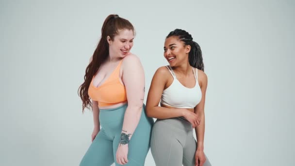 两名身穿运动服的跨国女孩背靠背地站在工作室灰色的背景下笑着 — 图库视频影像