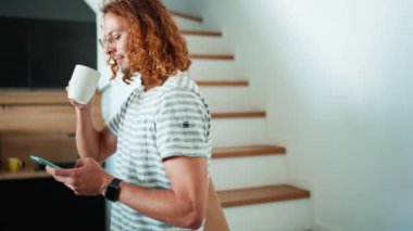 Gözlüklü neşeli kıvırcık saçlı adam bir fincan kahve içiyor ve ofis mutfağında cep telefonuna mesaj atıyor.