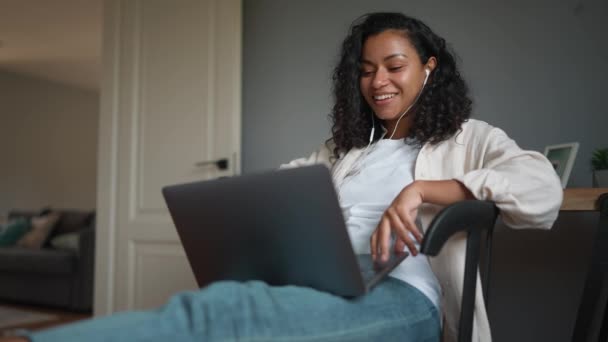 在家里用耳机和问候语在笔记本电脑上用视频交谈的卷曲的非洲女人笑着 远程工作 — 图库视频影像