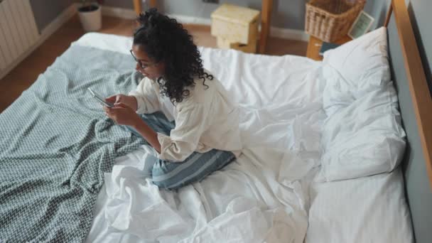 穿着眼镜的漂亮卷曲的非洲女人在家里的床上用手机发短信的头像 — 图库视频影像