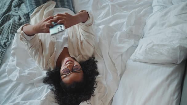 坐在眼镜上的快乐的卷曲的非洲女人在家里的床上用手机发短信的头像 — 图库视频影像