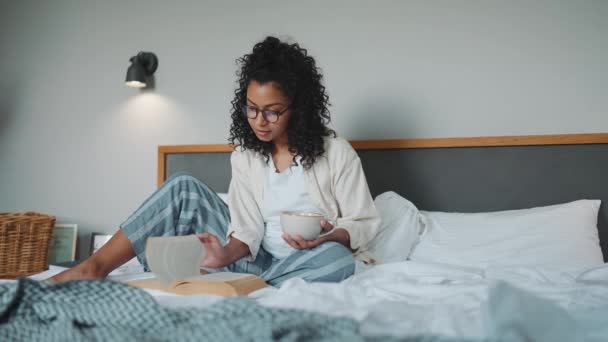 戴着眼镜的自信的卷曲的非洲女人在家里看书 床上放着一杯茶 — 图库视频影像