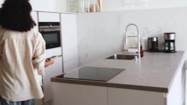 Gözlüklü komik Afrikalı kadın kulaklıkla müzik dinliyor ve evdeki buzdolabından yemek çıkarıyor.