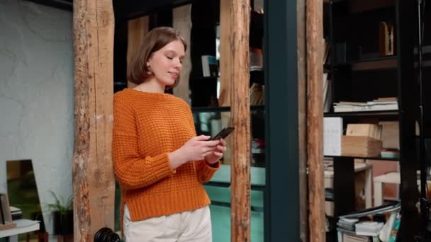 幸せな女性デザイナーを身に着けているオレンジ色のセーターテキストを携帯電話と周りを見回す共同作業スペース — ストック動画