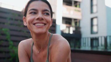 Cheerful brunette female athlete looking around and at camera before yoga practice on hotel balcony