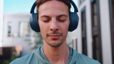 Close-up view of smiling curly haired sportsman opening eyes at camera and listening music in headphones on hotel balcony