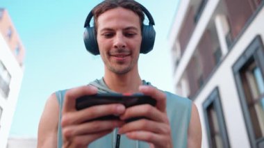 Pretty curly haired sportsman typing on smartphone and listening music in headphones on hotel balcony