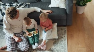 Kıvırcık saçlı sarışın baba, evde yerde kitap okurken oğluyla ve kızıyla konuşuyor.