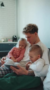 Ciddi kıvırcık saçlı, sarışın baba, kızını ve oğlunu kollarına alıp evdeki kanepede tablette çizgi film izliyor.