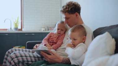 Yoğunlaşmış kıvırcık saçlı, sarışın baba, kızını ve oğlunu kollarına alıp evdeki kanepede tablette çizgi film izliyor.
