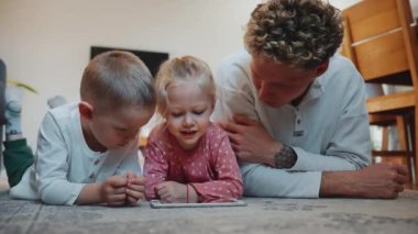 Mutlu kıvırcık saçlı genç baba evde yerde çocuklarla çizgi film izliyor.