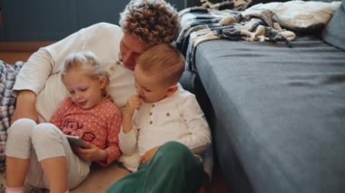 Güzel kıvırcık saçlı, sarışın, genç baba evde yerde çocuklarla çizgi film izliyor.