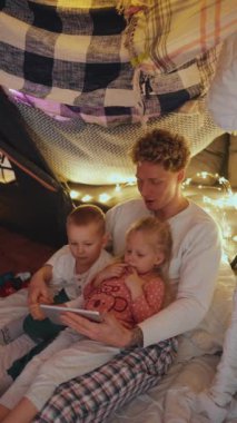 Gülen kıvırcık saçlı sarışın baba evde yatmadan önce oğluyla kızıyla tablette çizgi film izliyor.