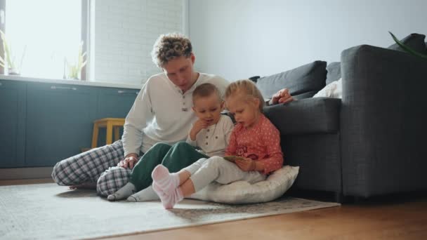 真剣な若いブロンドの娘は 自宅の床で父と弟に本を読んで — ストック動画
