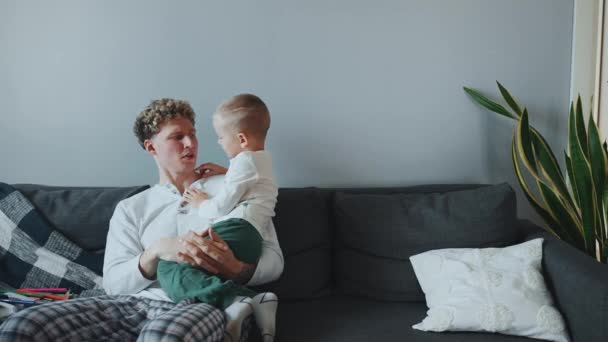 彼の腕に息子を抱え 自宅のソファーで彼に話しかけているカーリーヘアの若い父親 — ストック動画