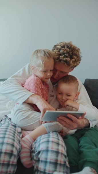 美しくかわいい髪のブロンドの父親は 自宅でソファーのタブレットで漫画を見ている彼の腕に娘と息子を抱いています — ストック動画