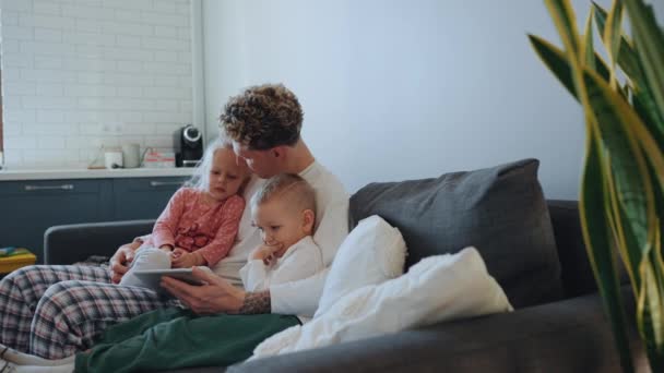 自宅でソファーのタブレットで漫画を見ている彼の腕に娘と息子を抱えている自信を持ってかわいい金髪の父親 — ストック動画