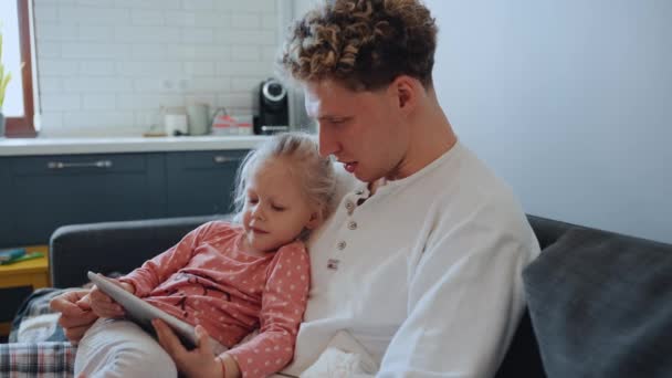 かわいいかわいい髪のブロンドの父親は 自宅でソファーのタブレットで漫画を見ながら娘を抱いています — ストック動画