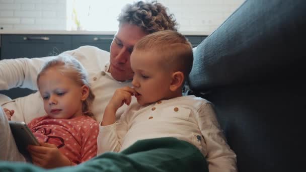 陽気なカーリーヘアブロンドの若い父親は子供と漫画を見て 自宅の床で息子を噛むために指を与える — ストック動画