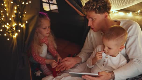笑顔のカーリーヘアブロンドの父親は 自宅で寝る前に息子と娘とタブレットで漫画を見ています — ストック動画