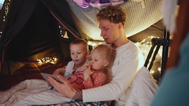 おかしいかわいい髪のブロンドの父親は 自宅で寝る前に息子と娘とタブレットで漫画を見ています — ストック動画