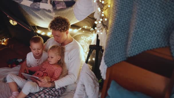 フレンドリーなカーリーヘアブロンドの父親は 自宅で寝る前に息子と娘とタブレットで漫画を見ています — ストック動画