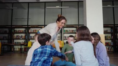 Gülümseyen öğretmen ve çocuklar avuçlarını bir araya getirip kütüphanede daireler çiziyorlar. Takım oluşturma kavramı