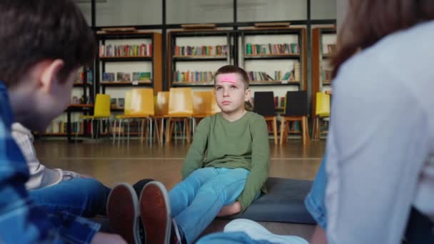図書館の床の円に座っている額の紙を持っているかを推測する真剣な子供たち — ストック動画