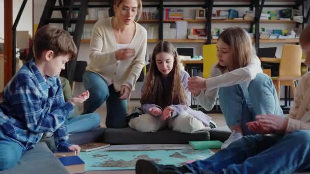 ポジティブな子供と教師がコインを保持し 図書館の床の世界地図を見る — ストック動画